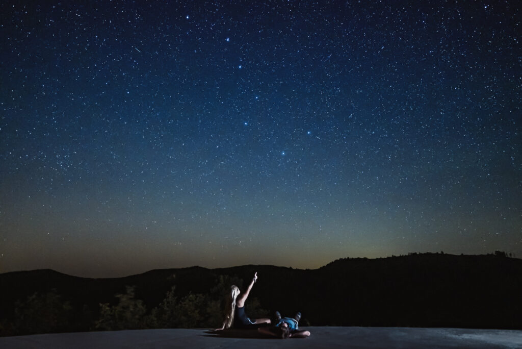 Two people lie on a surface, gazing up at a starry night sky.